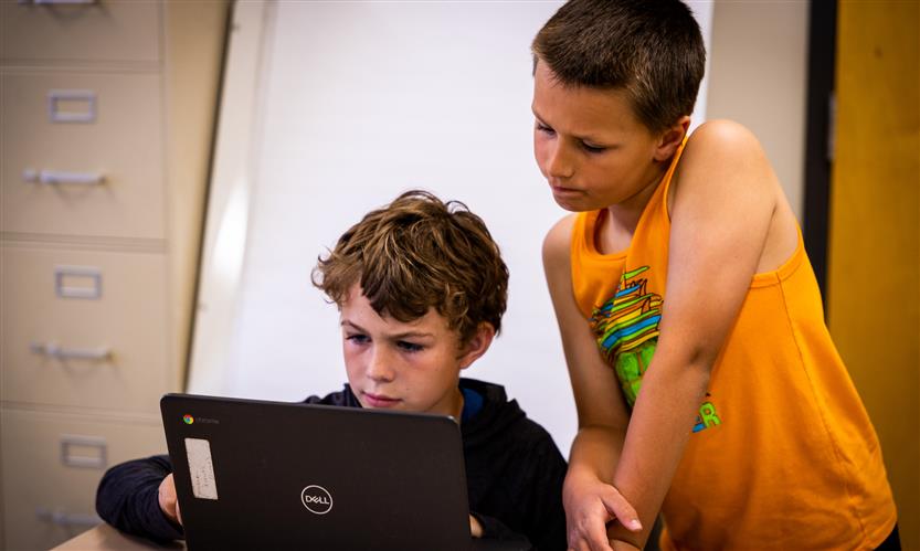 Two boys lean on a desk while using a Chromebook